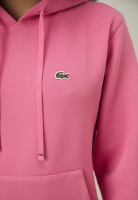 Sweat à capuche rose en tissu texturé avec une poche kangourou, arborant un petit logo crocodile vert Lacoste et des cordons de serrage.