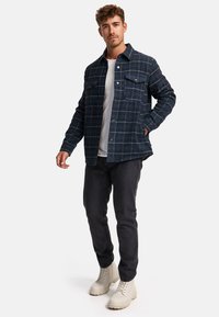 Navy flannel shirt jacket with a checked pattern, featuring two front pockets and button closure, paired with dark jeans and pale boots.