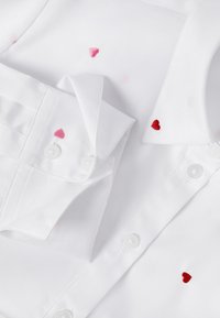 White button-up shirt with small embroidered red and pink hearts on collar, cuff, and chest areas, displayed flat.