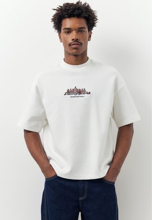 Muž s kučeravými vlasmi v bielom nadrozmernom tričku s grafikou malých postáv a francúzskym textom, tmavomodré rifle, ruky v kapsách