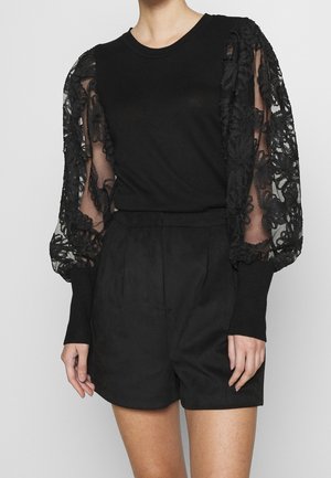 Personne vêtue d'une tenue noire avec des manches bouffantes en dentelle florale transparente et un short noir uni taille haute, sur un fond neutre.