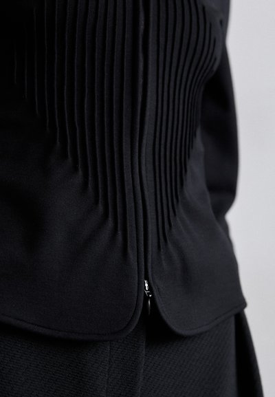 Čierny top s plisovanými detailmi, hladkou textúrou, zaobleným lemom a zipsovým zapínaním v spodnej časti. Detailný pohľad na dizajn látky.
