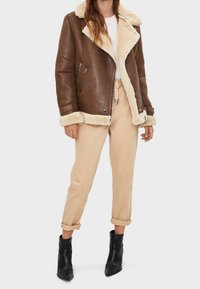 Giacca in shearling marrone con fodera in pelliccia beige, chiusura con zip e tasche laterali. Abbinata a pantaloni beige e stivali neri alla caviglia.