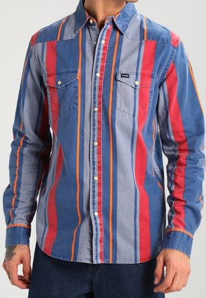 Mann trägt ein langärmliges Hemd mit Knöpfen, das vertikale rote, blaue, orange und graue Streifen, zwei Brusttaschen und ein kleines Logoetikett auf der linken Tasche aufweist.