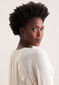 Blusa bianca plissettata con dettagli arricciati, indossata da una persona con capelli neri corti e texturizzati. Lo sfondo è neutro e discreto.