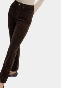 Braune Cordhosen mit geradem Bein, vorderen Taschen und einem Knopfverschluss, getragen mit einem hellen Oberteil und flachen Schuhen.