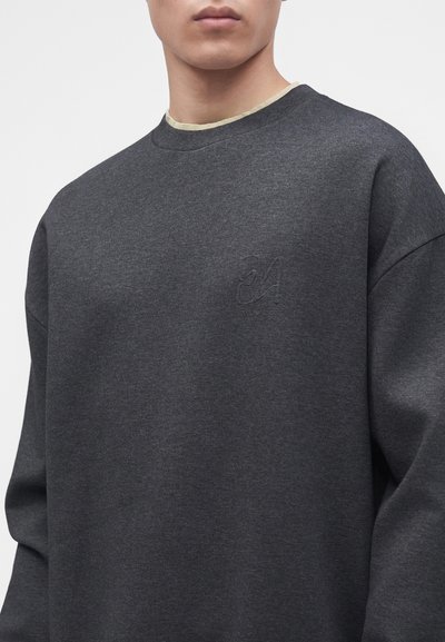 Homme portant un sweat-shirt gris foncé à col rond avec un logo ton sur ton discret sur la poitrine gauche, visible du cou jusqu'au milieu du torse.