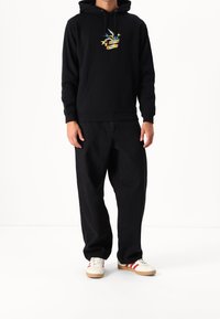 Svart huvtröja med framficka, prydd med en färgglad fågel och textdesign. Bärs med vida svarta byxor och vita skor med röda ränder.