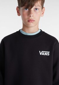 Sudadera negra con cuello redondo, que presenta un interior color azul claro y un logo blanco de Vans en la parte izquierda del pecho. Material suave, corte casual.