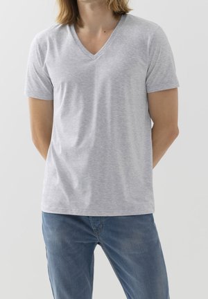 Osoba ubrana w jasnoszarą koszulkę z krótkim rękawem i dekoltem w serek oraz niebieskie dżinsy, stojąca z rękami za plecami na jednolitym jasnym tle.