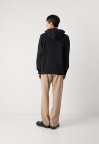 Sweat-shirt à capuche noir avec une coupe décontractée, associé à un pantalon beige clair, vu de dos sur un fond blanc.