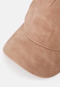 Căciulă de baseball din suede bej, cu o margine cusută și logo-ul embosat „Calvin Klein” pe partea din față. Textură moale cu linii curate.
