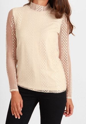 Femme portant un haut en dentelle beige à manches longues avec un col montant, associé à un pantalon noir, debout devant un fond uni.