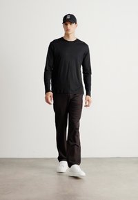 Ung mann som går fremover iført svart langermet skjorte, svarte bukser, svart caps med hvit tekst, og hvite sneakers mot en ensfarget bakgrunn.