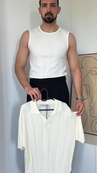 Koszulka polo z krótkim rękawem w kolorze złamanej bieli. Tekstura ribbed, kołnierzyk z wyłogami, pionowe paski i zapięcie na zamek z przodu. Wisi na czarnym wieszaku.
