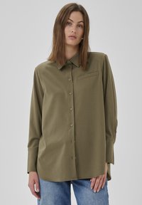 Frau mit glatten braunen Haaren, die ein olivgrünes, geknöpftes Langarmhemd und eine hellblaue Jeans trägt, vor einem schlichten grauen Hintergrund.