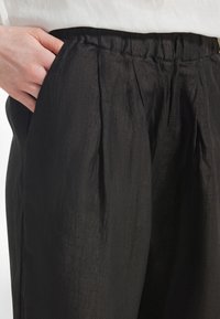 Hånd hviler i lommen på løse, sorte bukser med elastisk talje, båret med en hvid top.