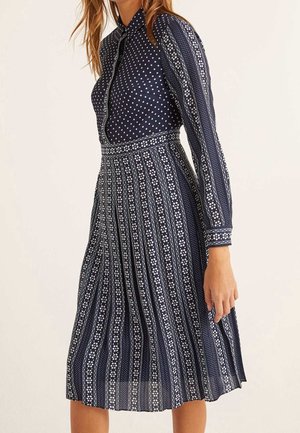 Nainen, joka käyttää laivastonsinistä mekkoa, jossa on valkoisia pilkku- ja raidallisia kuvioita, pitkät hihat ja pliseerattu midihame.