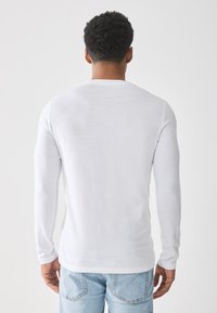 Camisa blanca de manga larga hecha de tela suave, con un cuello redondo y un diseño ajustado, combinada con jeans de color azul claro.