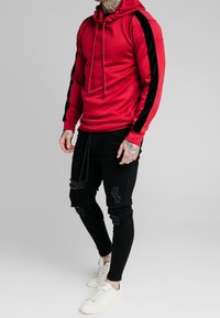 Felpa rossa con cappuccio e pannelli laterali neri, maniche lunghe e cordini, abbinata a jeans skinny neri strappati e scarpe da ginnastica bianche.