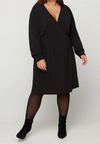 Czarna sukienka-wrap w miękkim materiale, z głębokim dekoltem w kształcie litery V, długimi rękawami i rozkloszowanym krojem. Noszona z czarnymi botkami do kostek.