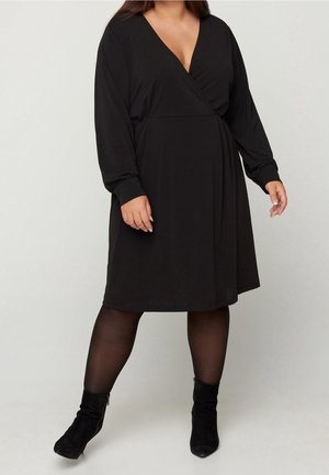 Robe noire enveloppante en tissu doux, avec un décolleté en V profond, des manches longues et une silhouette évasée. Portée avec des bottes noires à hauteur de cheville.
