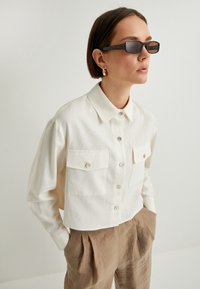 Camisa de botón recortada de un beige claro con dos bolsillos en el pecho, hecha de una tela suave, combinada con pantalones con pliegues beige y gafas de sol oscuras.