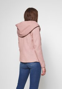 Lichtroze jack met een ontspannen capuchon, gestructureerd ontwerp en een textuurstof. Draagt met blauwe jeans, met details en zakken aan de achterkant.