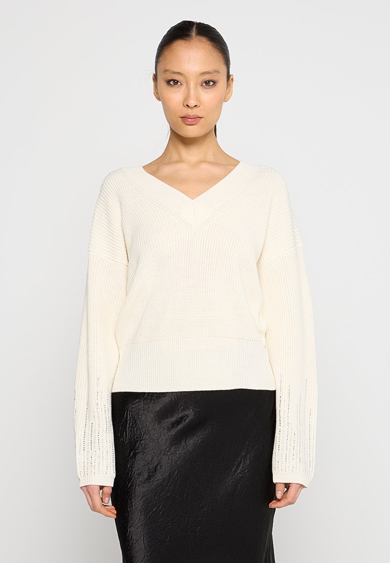 Maglione a V in maglia di colore crema con spalle cadenti, texture a coste e maniche ampie, abbinato a una gonna nera lucida fino al ginocchio.