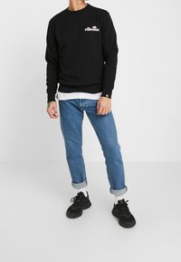 Felpa nera con scollo a girocollo e logo bianco, indossata sopra una camicia bianca, abbinata a jeans blu risvoltati e sneakers nere.