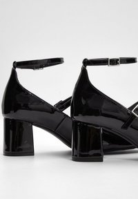 Scarpe con tacco alto in vernice nera, dotate di una cinghia alla caviglia e tacco quadrato. Presentano una punta affilata e un design a strisce incrociate.