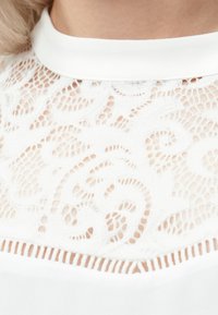 Κοντινής λήψης από λευκό υφασμάτινο δαντέλας με λουλουδάτα μοτίβα σε ρούχο με υψηλό λαιμό, το οποίο φοράει ένα άτομο.