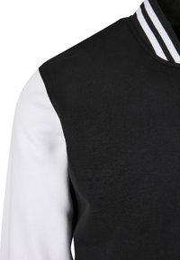 Veste universitaire noire avec manches blanches et col côtelé rayé noir et blanc, montrant le torse supérieur et la zone des épaules.