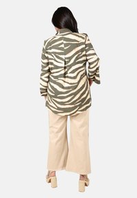 Femme portant une chemise à rayures zèbre vertes et beiges à manches longues, un pantalon large beige et des sandales à talons carrés beiges, de dos visible.