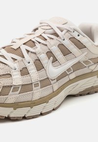 Beige atletiska skor med övre i mesh och mocka, vita accenter och en texturerad gummisula. Har förstärkt sömnad och en logotyp.