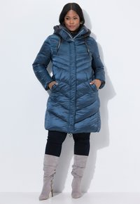 Giacca blu imbottita con design a chevron, cappuccio foderato in pelliccia sintetica, chiusura con zip e tasche laterali, indossata sopra pantaloni neri e stivali grigi.