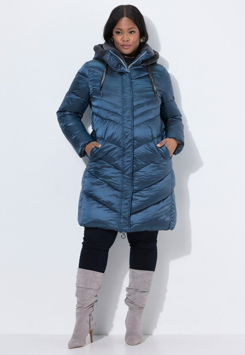 Giacca blu imbottita con design a chevron, cappuccio foderato in pelliccia sintetica, chiusura con zip e tasche laterali, indossata sopra pantaloni neri e stivali grigi.