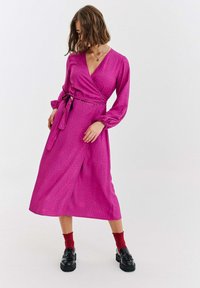 Fuksjowa sukienka kopertowa z długimi rękawami, zdobiona subtelnym wzorem w kropki, z wiązaniem w talii i długością do połowy łydki. Noszona z czarnymi butami i czerwonymi skarpetkami.
