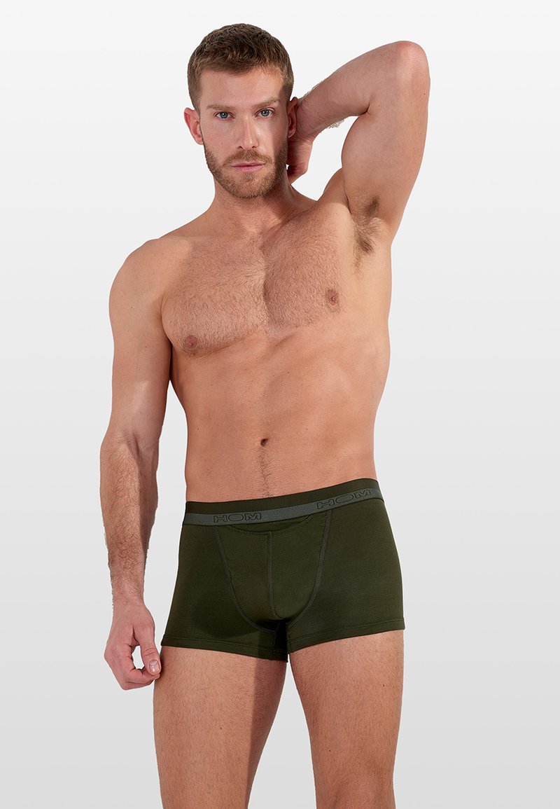 Hombre sin camiseta con cabello corto, vistiendo calzoncillos tipo bóxer color verde oscuro, levantando un brazo detrás de la cabeza contra un fondo claro y liso.