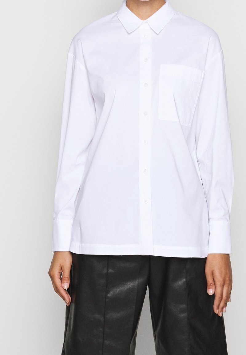 Camicia bianca con colletto a punta, maniche lunghe e taschino sul petto. Tessuto morbido, taglio dritto e pantaloni neri a contrasto.