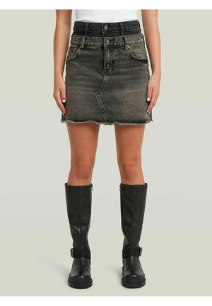 Frau trägt einen schwarzen Denim-Minirock mit doppeltem Bund und rohem Saum sowie schwarze kniehohe Lederstiefel, steht vor einem schlichten Hintergrund.