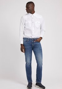 Camisa de botones blanca de manga larga con dos bolsillos en el pecho, combinada con jeans de mezclilla azules y zapatillas oscuras, de pie contra un fondo liso.