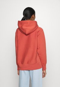 Hoodie w kolorze rdzy, wykonany z miękkiego materiału, z kieszenią typu kangur i prążkowanymi mankietami oraz dolnym brzegiem. Stylizowany z luźnymi, jasnymi dżinsami.