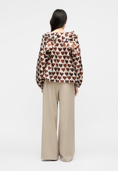 Γυναίκα με την πλάτη, που φοράει μπλούζα μακρυμάνικη με καφέ σχέδια καρδιές και μπεζ παντελόνι με φαρδιά πόδια σε απλό φόντο.