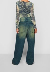 Halenka s dlouhým rukávem, průhledná, s vícebarevným vzorem a nápisem, v kombinaci s volnými, vybledlými modro-zelenými širokými džíny a výraznými zadními kapsami.