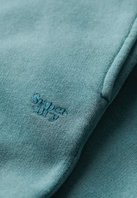 Tissu de sweat-shirt vert bleu clair avec une texture fine ; présente un logo "Superdry" brodé dans une teinte assortie. Design simple, sans motifs.