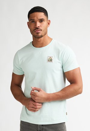 Homme aux cheveux courts et à la barbe portant un t-shirt bleu clair avec une petite écusson floral sur la poitrine, debout devant un fond uni.