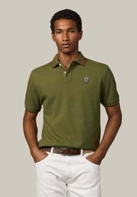Olivovo zelené polo tričko s červenými akcentmi na golieri a logo nášivkou, krátke rukávy a klasický strih, skombinované s bielymi nohavicami a hnedým opaskom.