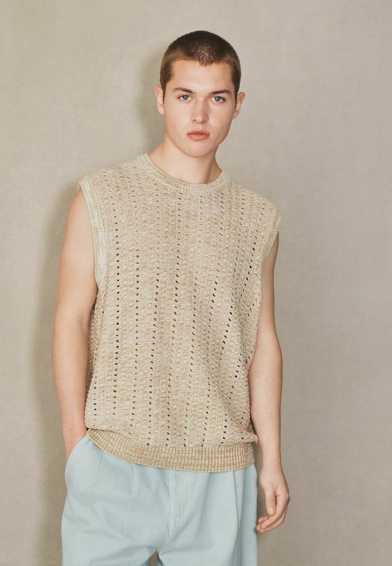 Ung mand med kort hår iført en ærmeløs beige strikket top og lyseblå bukser, stående mod en enkel neutral baggrund.