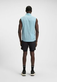 Light blue sleeveless vest made from a smooth, lightweight fabric, featuring a rear pocket and a streamlined design, worn with black shorts.